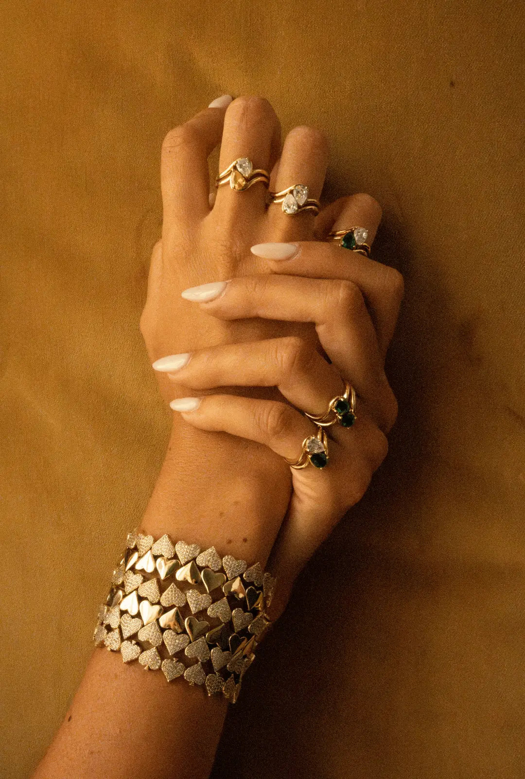 Mains croisées portant des bagues en plaqué or décorées de pierres en zirconium coupe poire et des bracelets en plaqué or en forme de multiple coeur, dont certains sont pavés de zirconiums.