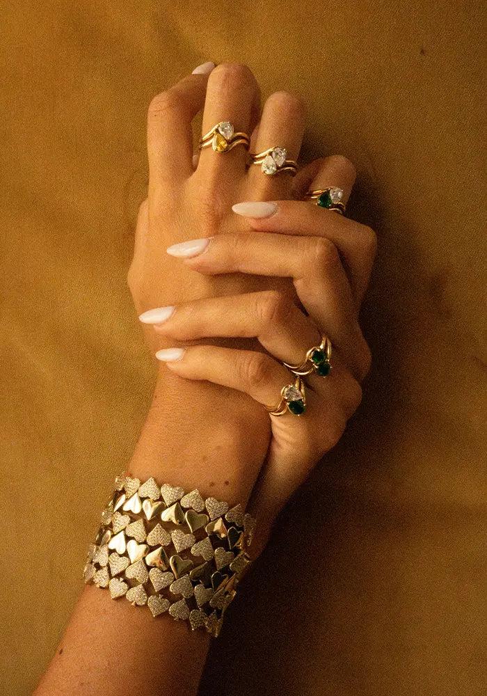 Mains croisées portant des bagues en plaqué or décorées de pierres en zirconium coupe poire et des bracelets en plaqué or en forme de multiple coeur, dont certains sont pavés de zirconiums. 
